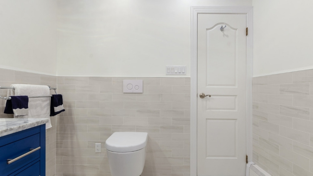 Μικρό μπάνιο με κρεμαστή λευκή λεκάνη τουαλέτας και πόρτα και μπλε συρταριέρα.
