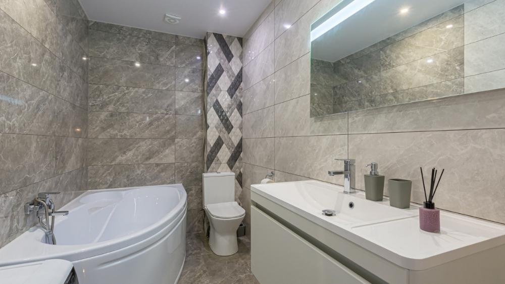 Στενόμακρο μπάνιο με λευκό νιπτήρα, μπανιέρα, λεκάνη και πλακάκια με γκρι αποχρώσεις.