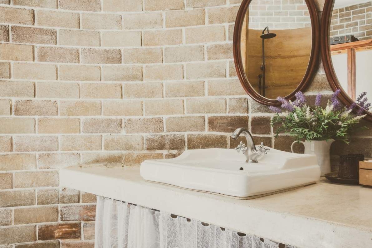 Λευκός νιπτήρας από παλιό μπάνιο και διακόσμηση με καθρέπτη και φυτά σε φόντο τούβλινου τοίχου.