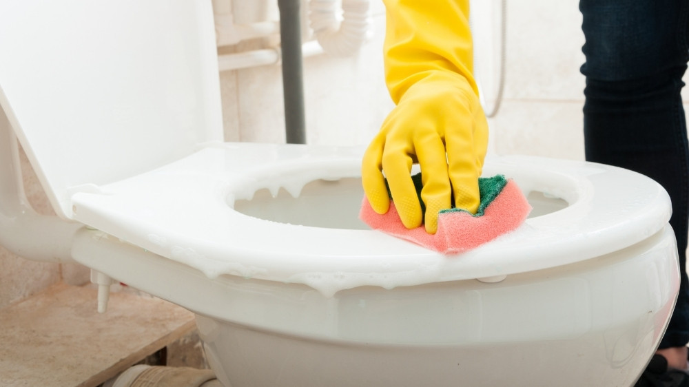 Χέρι με κίτρινο γάντι και ροζ-πράσινο σφουγγαράκι καθαρίζει λεκάνη τουαλέτας.