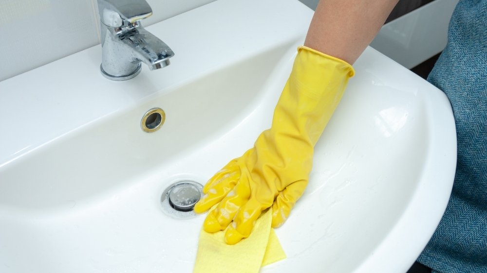Χέρι με κίτρινο γάντι καθαρίζει νεροχύτη μπάνιου με βετέξ. 