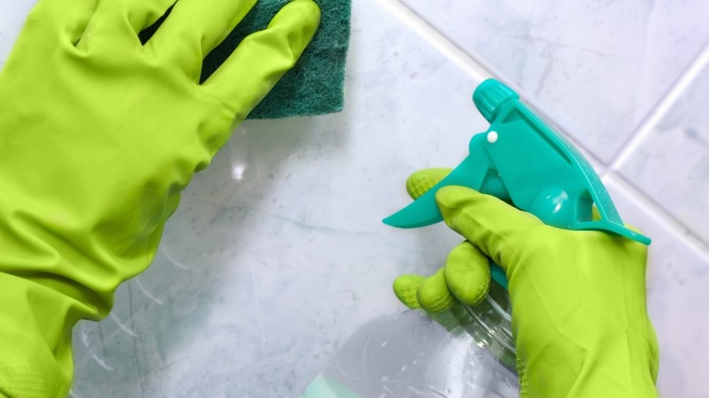 Χέρια με πράσινα γάντια, σφουγγαράκι και απολυμαντικό σπρέι καθαρίζουν τοίχους μπάνιου.