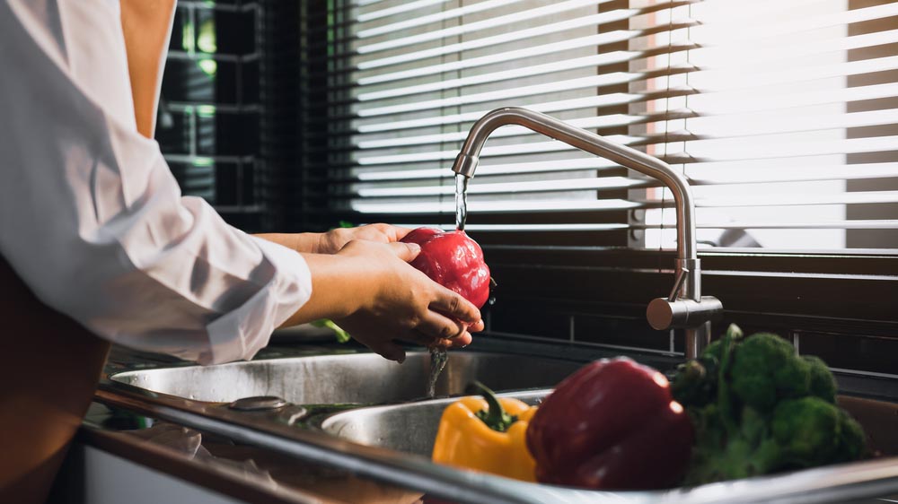 Άνθρωπος με ποδιά πλένει λαχανικά στη βρύση της κουζίνας, ενώ εισέρχεται από το παράθυρο το φως της μέρας.