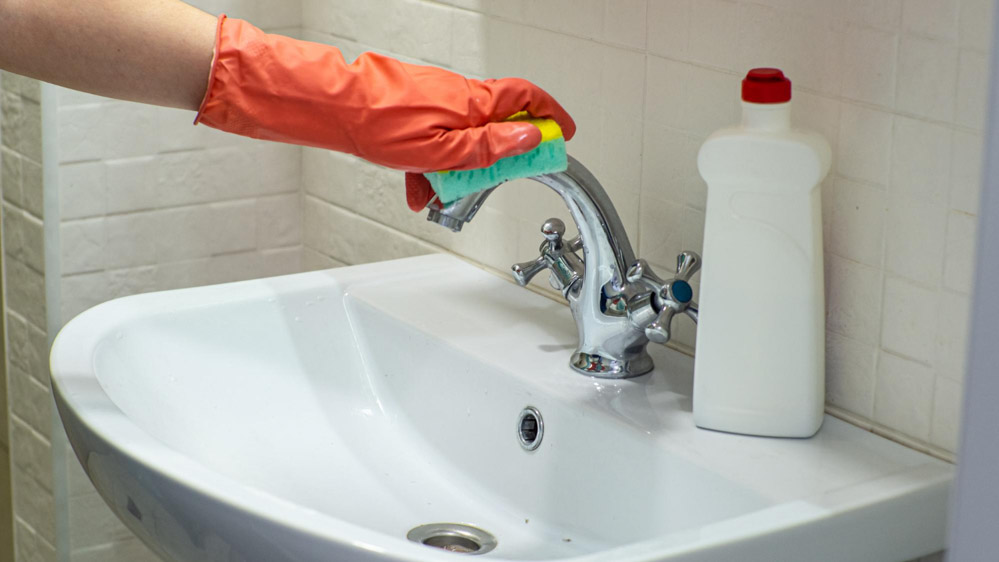 Χέρι με πορτοκαλί γάντι καθαρίζει μια βρύση μπάνιου από άλατα με σφουγγάρι.