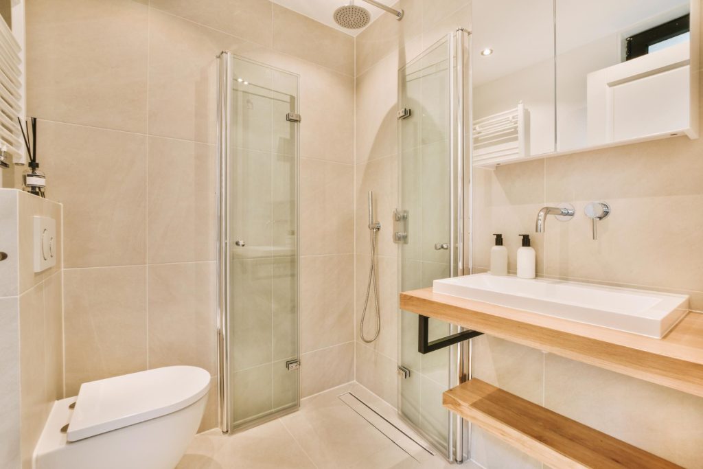 Μοντέρνα χτιστή ντουζιέρα σε μικρό μπάνιο με ανοιγόμενες γυάλινες πόρτες, ανάμεσα σε λεκάνη τουαλέτας και νιπτήρα.