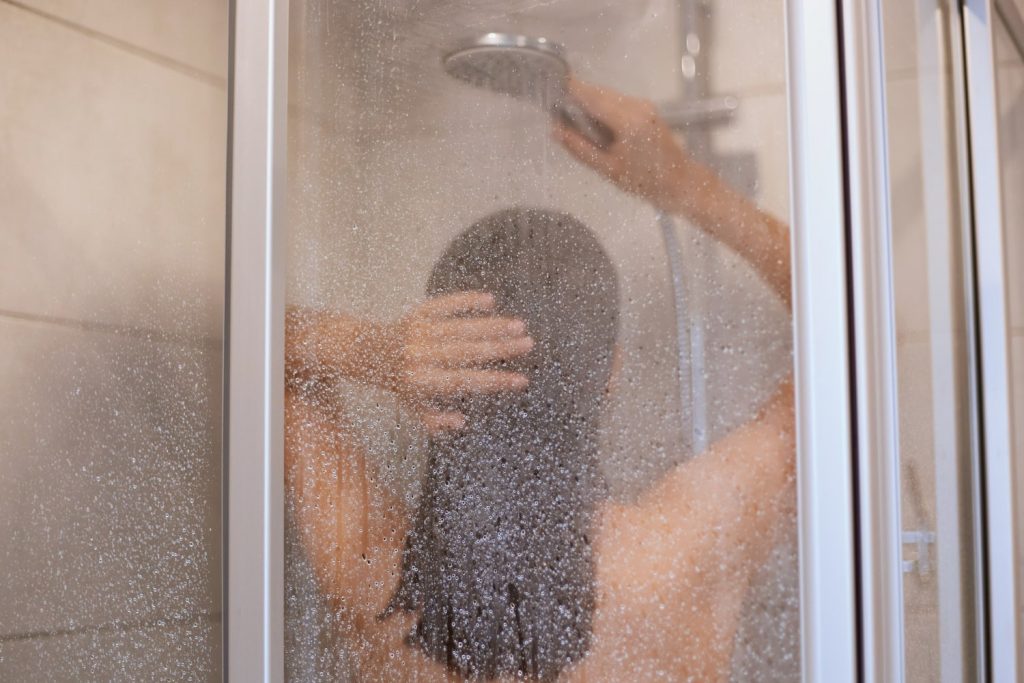 Θεματική εικόνα για το πώς καθαρίζω το τζάμι της ντουζιέρας. Γυναίκα κάνει ντουζ σε ντουζιέρα, το τζάμι της οποίας είναι θολό.